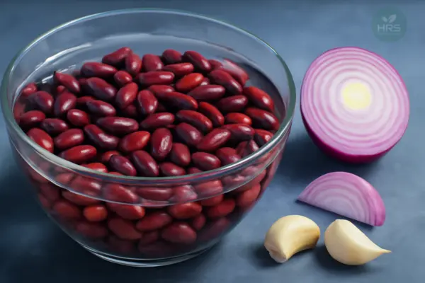 Raw kidney beans soaking in glass bowl with sliced onions and garlic cloves nearby.